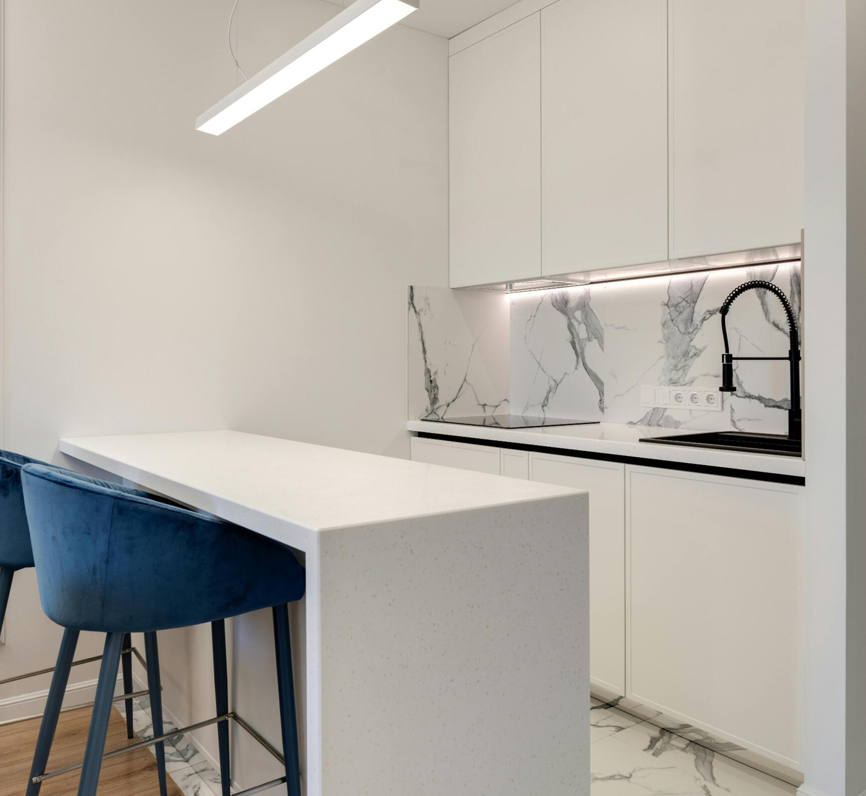 Modern white handleless kitchen with stone benchtops, black sink & mixer, and black glass induction cooktop A clean, minimalist kitchen featuring white handleless cabinetry, stone benchtops, a black pull‑out mixer, a black induction cooktop, and blue velvet bar stools. This modern layout reflects the simple, elegant renovation style offered by Sydney Budget Kitc